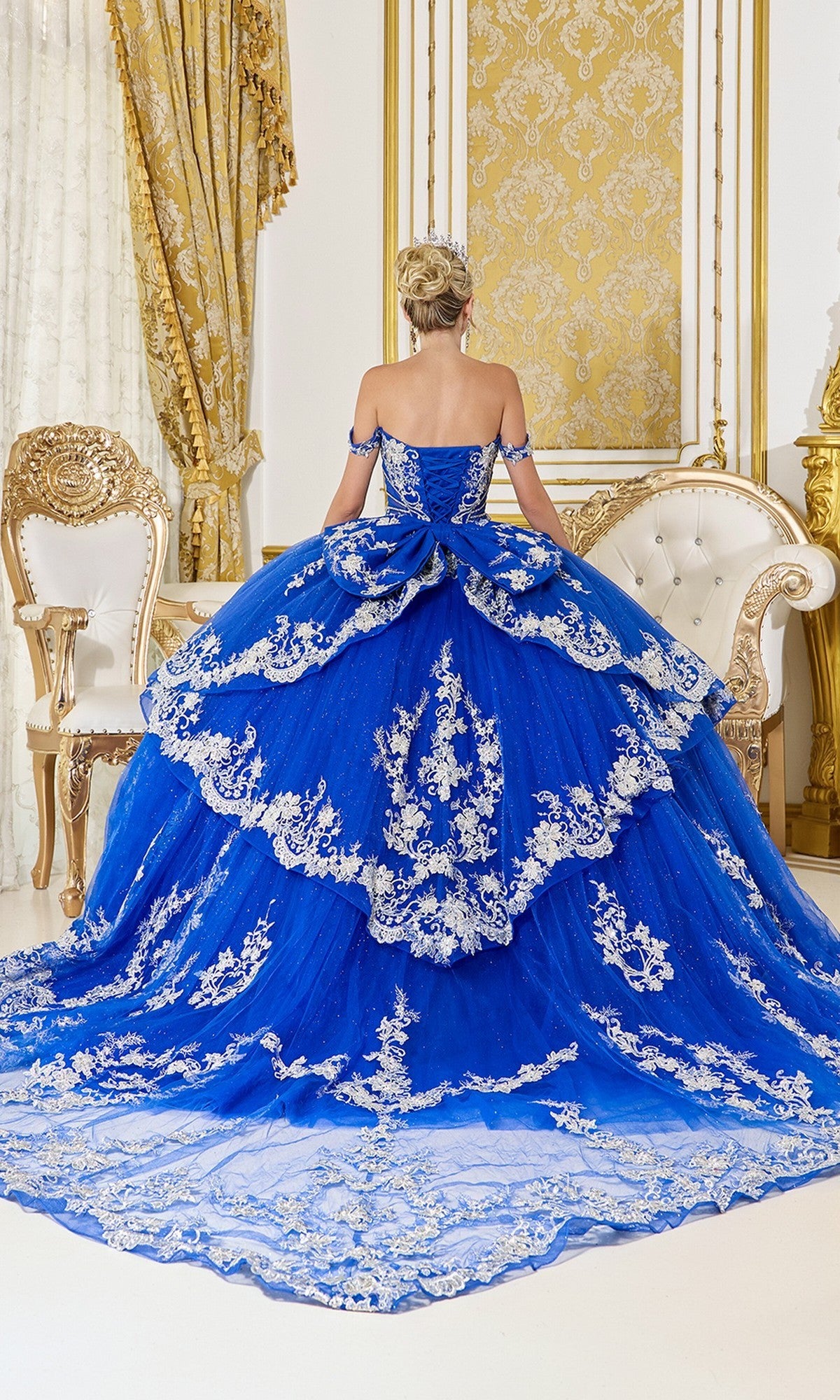 A woman wearing the Quinceanera Dress 5142 By Dancing Queen, featuring a glitter tulle skirt and corset bodice in royal blue, stands facing away in an ornate, gold-accented room with a white and gold sofa and chair.